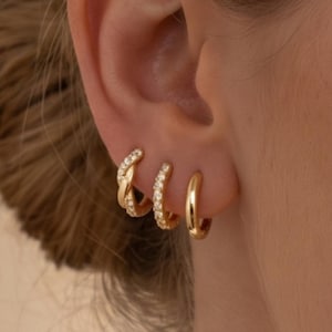 May include: Three gold hoop earrings worn in a row on an earlobe. The first earring is a twisted gold hoop with small clear stones, the second is a thin gold hoop with clear stones, and the third is a plain, smooth, polished gold hoop earring.