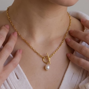 Peut inclure: Un collier à chaîne dorée doté d'un fermoir à bascule et d'un pendentif unique en perle blanche de forme irrégulière. Le collier est composé de maillons ovales et repose sur la clavicule, mettant en valeur une finition métallique.