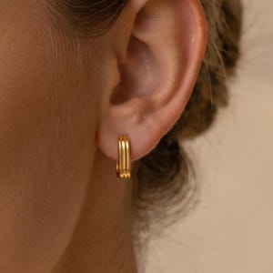 Pode incluir: Um pequeno brinco de argola dourado com um design de banda tripla. O brinco tem um acabamento metálico polido e uma forma arredondada e contínua que se ajusta ao lóbulo da orelha, exibindo uma aparência em camadas com três cristas paralelas.