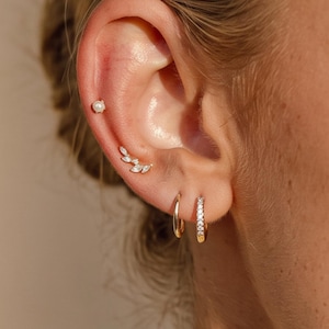 Könnte beinhalten: Ein goldenes Ohr-Set bestehend aus einem kleinen weißen Perlenstecker, einem geschwungenen goldenen Climber-Ohrring mit klaren Steinen im Marquise-Schliff, einem schlichten goldenen Creolen-Ohrring und einer goldenen Creole mit kleinen klaren Steinen.