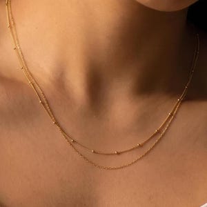 Könnte beinhalten: Eine zarte, zweireihige Goldkette mit einer feinen Kette und kleinen, gleichmäßig verteilten Goldperlen entlang der Stränge. Die Kette liegt am Schlüsselbein an und zeigt ein schlichtes Design mit zwei unterschiedlichen Kettenlängen.