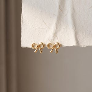 May include: A pair of gold-toned stud earrings shaped like small bows. Each earring features a smooth, polished metallic finish with a central knot and two symmetrical loops extending outward, creating a classic bow silhouette for the ears.