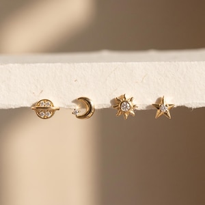 Puede incluir: Un conjunto de cuatro pendientes de botón en tono dorado con motivos celestiales. Los diseños incluyen un planeta con anillo, una luna creciente, un sol con puntas radiantes y una estrella de cinco puntas. Cada pendiente tiene pequeñas piedras claras.