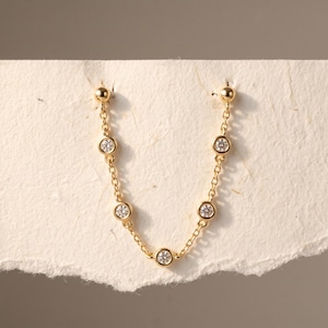 May include: A gold chain earring featuring five bezel-set round clear stones spaced along the length of the chain. The earring has a small gold ball stud at each end, creating a draped U-shape design that connects two points on a textured paper surface.