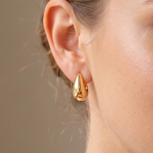 May include: A polished gold hoop earring in a smooth, teardrop shape. The earring features a reflective, metallic surface with a rounded, bulbous design that tapers to a point at the top, securely fastened through the earlobe with a seamless finish.