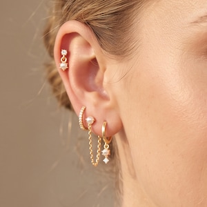 Op de afbeelding: Een set goudkleurige oorbellen met een kleine knop met een heldere steen en parelhanger, een pavé-ring, een parelknop verbonden door een fijne gouden ketting aan een tweede parelknop, en een gouden ring met een parel en heldere steen hanger.