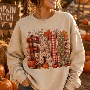 Op de afbeelding: Een beige sweater met ronde hals en een grote grafische print van het woord FALL. Elke letter heeft een uniek patroon, waaronder rood ruitjesmotief, bloemen, rood vichy-ruitje en groene stippen, omringd door illustraties van pompoenen en herfstbladeren.