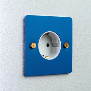 Puede incluir: Una placa de cubierta cuadrada azul para enchufe eléctrico con un centro circular blanco y dos pequeños tornillos de color latón situados a los lados izquierdo y derecho de la abertura central, montada sobre una pared blanca texturizada.