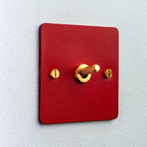 Op de afbeelding: Een vierkante rode lichtschakelaar met een getextureerde matte afwerking. De schakelaar is voorzien van een centrale goudkleurige metalen tuimelschakelaar met een gekartelde grip en twee bijpassende goudkleurige platte schroeven.