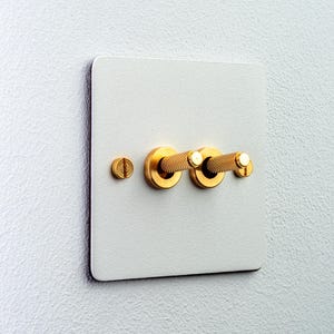 Könnte beinhalten: Eine quadratische weiße Lichtschalterplatte mit zwei goldenen zylindrischen Kippschaltern mit strukturierter Rändelung. Die Platte ist mit zwei goldenen Schlitzschrauben befestigt, die sich ganz links und ganz rechts an der Schaltereinheit befinden.