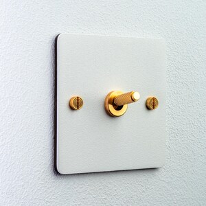 Pode incluir: Um interruptor de luz quadrado com uma placa frontal branca texturizada e uma alavanca central dourada. A alavanca apresenta um padrão de aderência serrilhado. Dois parafusos dourados de cabeça chata estão posicionados horizontalmente.