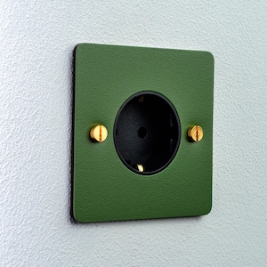 Può includere: Una presa elettrica quadrata con una placca frontale verde bosco opaco, un interno circolare nero e due viti in ottone lucido posizionate sui lati sinistro e destro dell'apertura centrale, montata a filo su una parete strutturata.