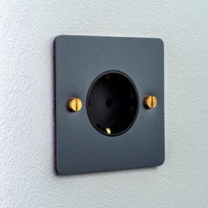 Op de afbeelding: Een vierkant, donkergrijs elektrisch stopcontact met een matte afwerking. Het stopcontact heeft een cirkelvormig zwart middenstuk en is aan de muur bevestigd met twee opvallende, platte messing schroeven aan de linker- en rechterkant.