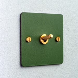 Pode incluir: Um interruptor de luz quadrado com um acabamento verde-floresta mate e texturizado. O interruptor apresenta uma alavanca central de metal dourado com textura serrilhada e dois parafusos de cabeça chata dourados em cada lado.