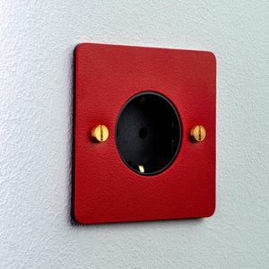 Op de afbeelding: Een vierkant stopcontact met een textuurrijke rode afdekplaat en een zwart rond middenstuk. De afdekplaat is bevestigd met twee platte messing schroeven aan de linker- en rechterkant van de centrale opening van het stopcontact.
