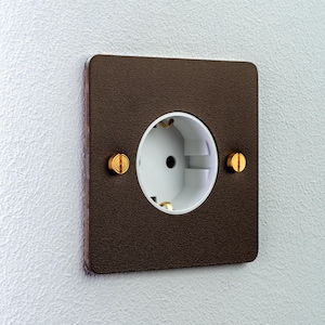May include: A square electrical wall socket featuring a dark brown textured faceplate with rounded corners. The centre contains a white circular socket insert, secured by two prominent gold-coloured flathead screws on the left and right sides.
