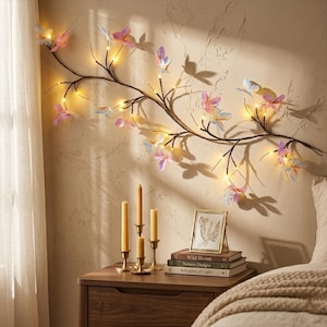 Pode incluir: Um ramo decorativo de parede com luzes LED brancas quentes e vários ornamentos de borboletas em tons pastel, incluindo rosa, azul e roxo, dispostos horizontalmente sobre uma mesa de cabeceira de madeira com velas.