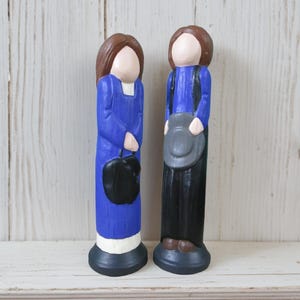 May include: Two hand-painted wooden peg dolls standing side by side. The left figure wears a blue dress with a white hem and holds a black bag. The right figure wears a blue shirt, black trousers, and holds a grey hat. Both have brown hair.