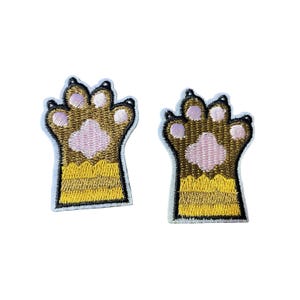 May include: Two embroidered patches shaped like animal paws. Each patch features a brown upper section with light pink paw pads and a yellow lower section with horizontal stripes, all outlined in black thread against a white fabric border.