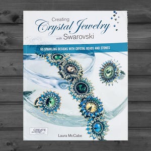 May include: A softcover book titled Creating Crystal Jewelry with Swarovski by Laura McCabe. The cover features a blue and green beaded bracelet and two matching rings, with the subtitle 65 sparkling designs with crystal beads and stones.