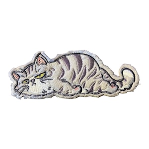 May include: An embroidered patch featuring a light cream-colored cat with dark gray tabby stripes lying down in a relaxed position. The cat has yellow eyes, a grumpy facial expression, and a curled tail, with visible stitching throughout the design.