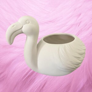 May include: A matte white ceramic planter shaped like a flamingo. The bird figure features a curved neck, a distinct beak, and textured wing details on the body. The top of the flamingo's back has a hollow, circular opening for holding items.