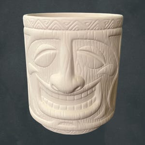 May include: A matte white ceramic tiki mug featuring a sculpted face with a wide, toothy grin, closed eyes, and a prominent nose. The rim is decorated with a repeating triangular geometric pattern, and the surface has a subtle wood grain texture.