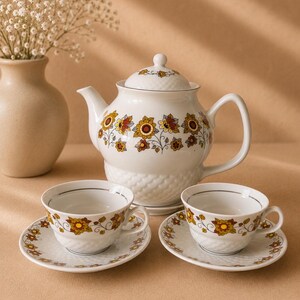 May include: A white porcelain tea set comprising a teapot with a lid and two matching teacups with saucers. Each piece is decorated with a floral pattern in shades of yellow, orange, and brown, and includes a textured basket-weave base design.