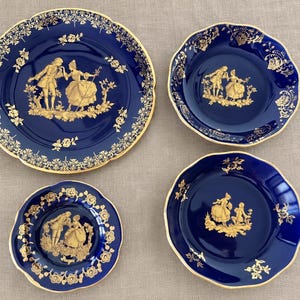 Può includere: Un set di quattro piatti in porcellana blu profondo con bordi smerlati e finiture dorate. Ogni piatto presenta un'illustrazione centrale dorata di due figure in abiti storici, circondata da intricati motivi floreali dorati lungo i bordi.