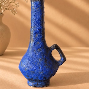 May include: A tall, narrow-necked ceramic vase featuring a vibrant cobalt blue glaze with a heavily textured, cratered surface and dark brown undertones. The vessel has a rounded base, a single integrated handle, and the text MyVintageHeritage.