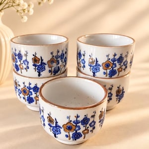 May include: A set of four white ceramic tea cups featuring a hand-painted floral pattern in shades of blue and metallic gold. Each cup has a smooth, rounded shape with a thin gold-toned rim. The text MyVintageHeritage is visible at the bottom right.
