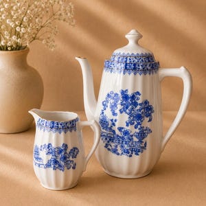 May include: A white porcelain teapot and matching creamer pitcher featuring a blue floral pattern, vertical fluted detailing, and gold trim accents. The set includes a lidded teapot with a curved handle and spout, and a small pouring pitcher.