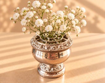 May include: A silver-toned metal flower frog vase with a decorative embossed rim and base, containing a cluster of small white baby's breath flowers. The text MyVintageHeritage is printed in the bottom right corner of the image.