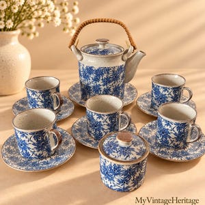 May include: A ceramic tea set featuring a teapot with a woven handle, five cups with matching saucers, and a lidded sugar bowl. Each piece is off-white with a speckled finish and a blue floral pattern. The text MyVintageHeritage is visible.