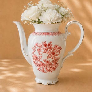 May include: A white porcelain teapot featuring a red transferware floral pattern and gold trim, filled with a bouquet of white roses and baby's breath. The teapot has a curved handle, a long spout, and a scalloped base with decorative detailing.