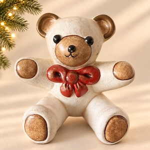 May include: A ceramic teddy bear figurine featuring a cream-colored body with brown accents on the ears, snout, paws, and feet. The bear has black eyes and a small black nose, and it wears a prominent red bow tied around its neck area.