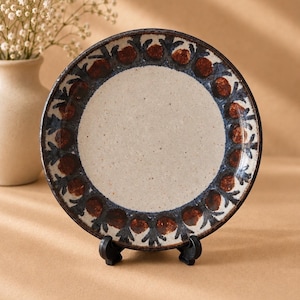 May include: A round ceramic plate featuring a speckled cream-colored center and a decorative border. The rim displays a repeating pattern of dark blue leaf-like shapes interspersed with circular reddish-brown elements on a light background.