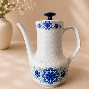 Puede incluir: Una cafetera de porcelana blanca con un patrón de diamantes texturizado en la parte superior y un motivo floral azul y gris vibrante alrededor de la base y la tapa. La cafetera incluye un asa curva, un pico largo y un pomo de tapa azul oscuro.
