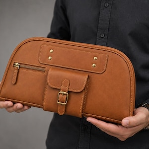 Puede incluir: Un bolso de mano de cuero color canela con borde superior semicircular, que presenta un bolsillo lateral con cremallera, un bolsillo central con solapa y hebilla de latón, y cuatro remaches decorativos de latón en la parte superior y una anilla.