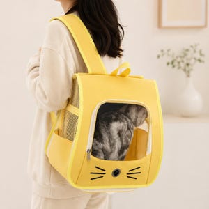 Puede incluir: Una mochila transportadora para mascotas de color amarillo brillante con una ventana transparente, paneles laterales de malla para ventilación y un diseño de cara de gato con bigotes y una pequeña ventilación circular.