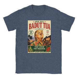 May include: A heather blue t-shirt featuring a graphic print of a person with clown makeup wearing a floral shirt. The text reads ARAK BALI BADUT TUA, SPIRIT TRADISIONAL, 40% VOL, 700 ml, and TOO LOUD FOR PARADISE, along with tropical palm tree imagery.