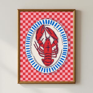 Op de afbeelding: Een ingelijste kunstdruk met een rode kreeft gecentreerd op een ovaal wit bord met een blauw gestreepte rand. De achtergrond bestaat uit een levendig rood en roze ruitpatroon. De lijst is goudkleurig met een dun profiel.