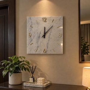 May include: A square wall clock with a white marble face featuring grey veining. The clock has gold-colored numerals at the 3, 6, 9, and 12 positions, with gold-colored stick markers for the other hours and black hands indicating the time.