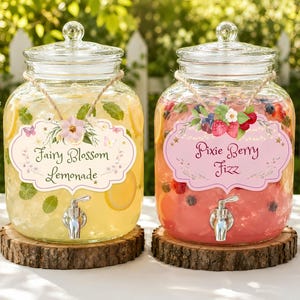 May include: Two clear glass beverage dispensers with lids and silver spigots, each resting on a round wood slice. The left dispenser contains yellow lemonade with mint and lemon slices, labelled Fairy Blossom Lemonade. The right contains red berry drink, labelled Pixie Berry Fizz.