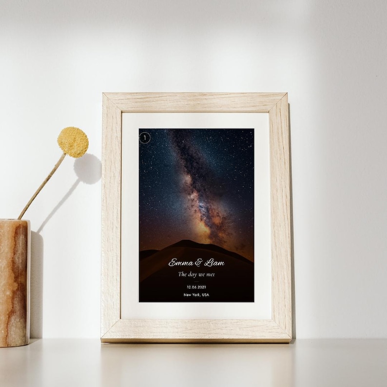 K&ouml;nnte beinhalten: Ein heller Holzrahmen mit einem vertikalen Druck eines Nachthimmels, der die Milchstra&szlig;e &uuml;ber einer dunklen Silhouette von Sandd&uuml;nen zeigt. Der Druck enth&auml;lt den Text Emma & Liam, The day we met, 12.06.2021 und New York, USA.