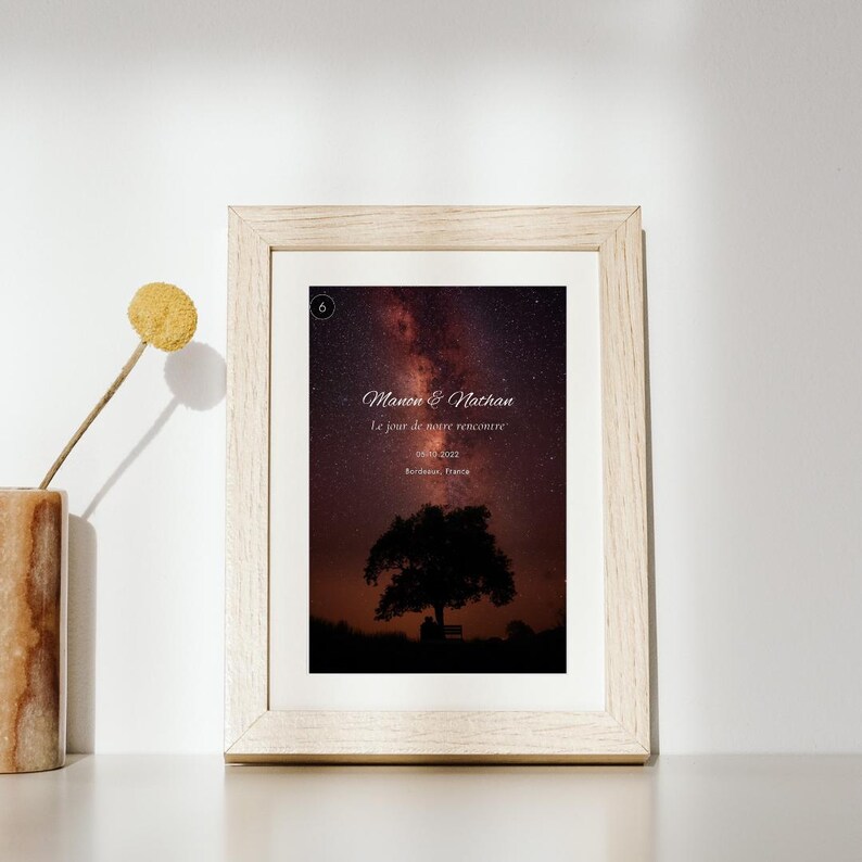 K&ouml;nnte beinhalten: Ein heller Holzrahmen mit einem Druck eines Nachthimmels mit einer sternenklaren Galaxie &uuml;ber der Silhouette eines Baumes und einer Bank. Der Text lautet Manon & Nathan, Le jour de notre rencontre, 05.10.2022, Bordeaux, France, mit einem kleinen Kreis, der eine 6 enth&auml;lt.