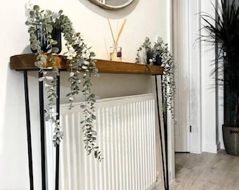 May include: A narrow wooden console table with thin black metal hairpin legs, positioned over a white radiator. The table features two black pots containing trailing green eucalyptus plants and a small glass diffuser with reed sticks.