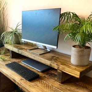 May include: A dark stained wooden monitor stand with a rectangular top and two vertical supports. The stand holds a black computer monitor, while a black keyboard and a closed laptop are positioned on the desk surface beneath the shelf.