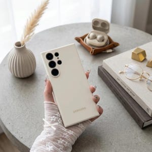 May include: A cream-colored smartphone case with a rectangular shape and a cutout for a multi-lens camera system on the back. The word SAMSUNG is printed in a subtle font near the bottom center of the smooth, matte-finish protective cover.
