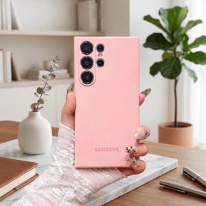 May include: A light pink smartphone case with a smooth matte finish, featuring a rectangular cutout for the camera array on the upper left side and the word SAMSUNG printed in a matching pink tone near the bottom center of the back panel.
